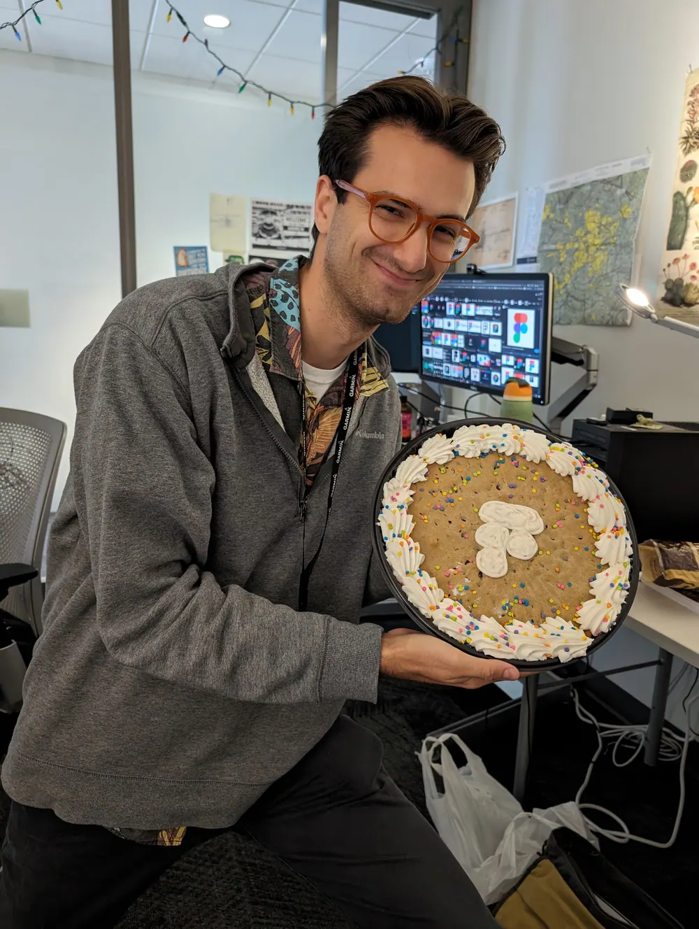 Andrew at his desk, grinning and holding up a giant cookie cake